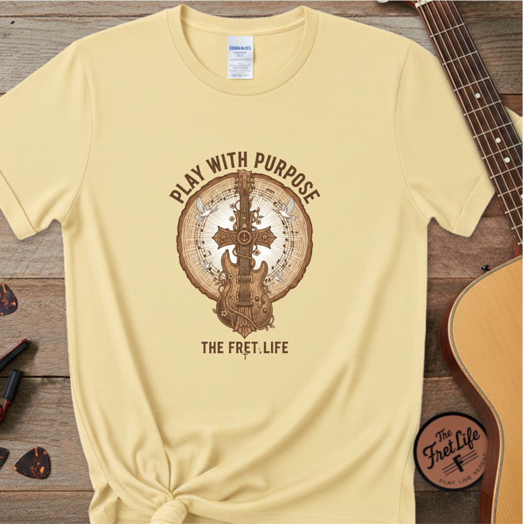 Beige t-shirt with guitar graphic and text 'Play with Purpose, The Fret Life' on a wooden surface with a guitar.