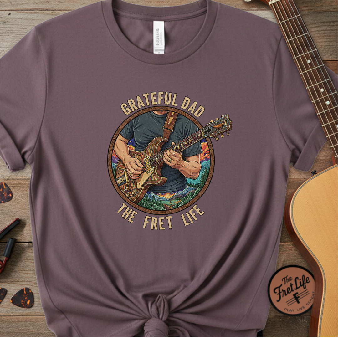 Purple t-shirt with 'Grateful Dad The Fret Life' graphic on a wooden surface with a guitar.