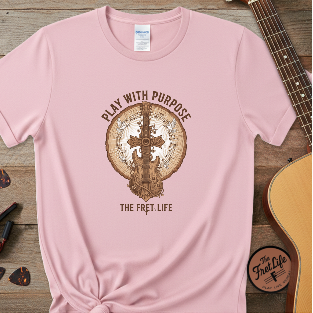 Pink t-shirt with guitar graphic and text 'Play with Purpose, The Fret Life' on a wooden surface with a guitar.