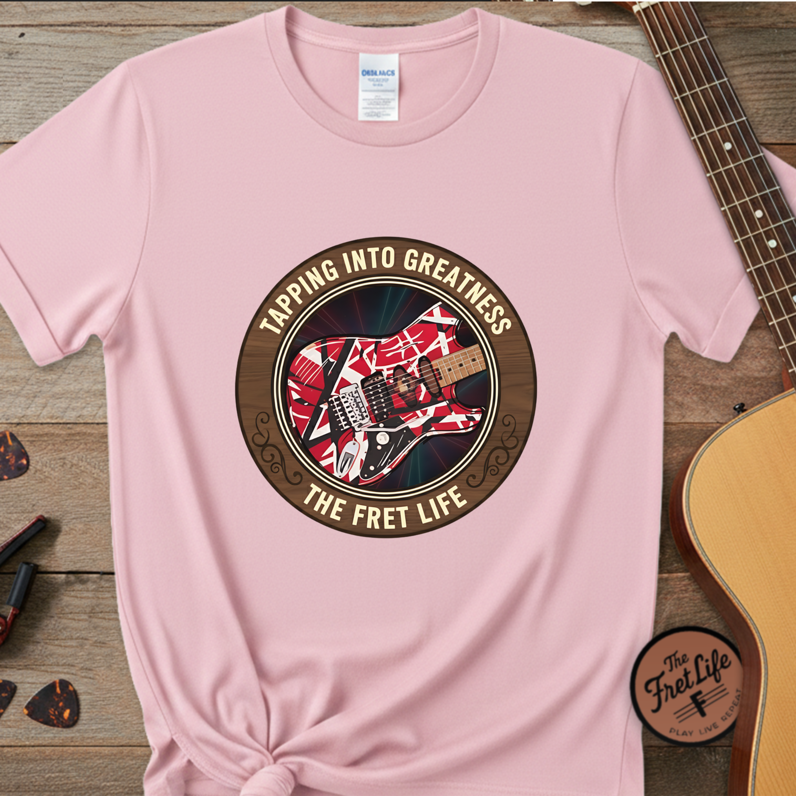 Pink t-shirt with guitar graphic and text 'Tapping into Greatness, The Fret Life' on a wooden surface with a guitar.