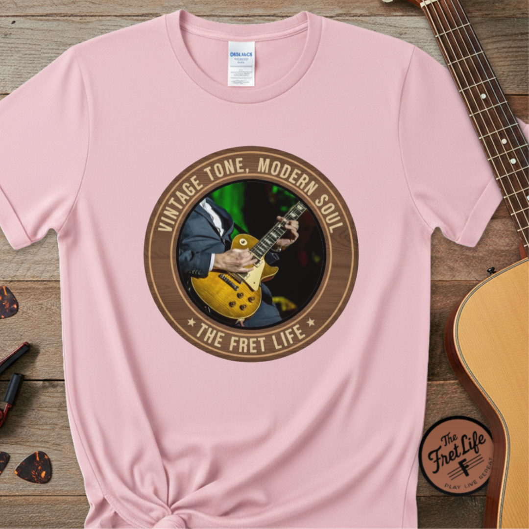 Pink t-shirt with guitar graphic and 'The Fret Life' text on a wooden surface with a guitar.