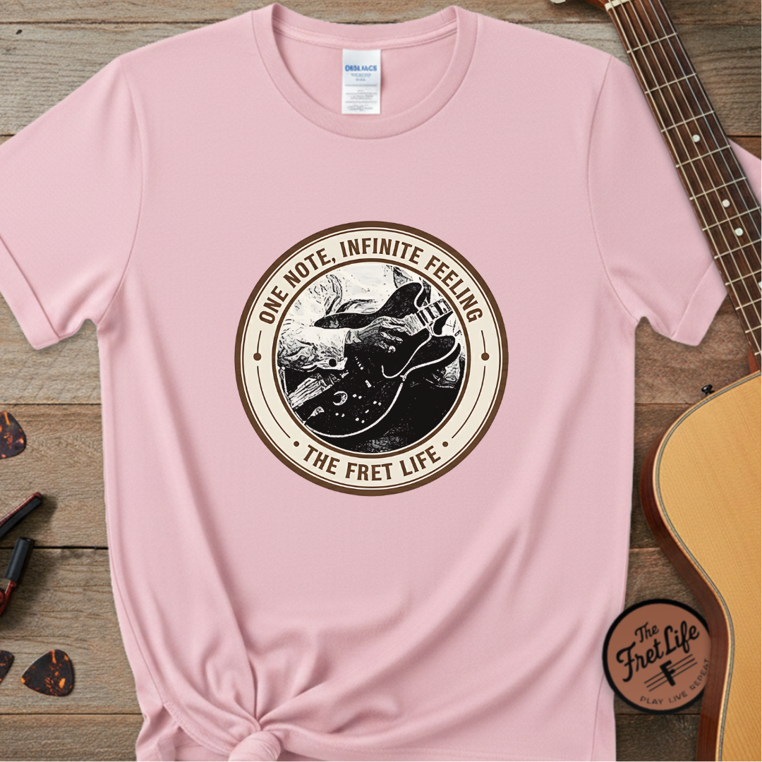 Pink t-shirt with a guitar graphic and text on a wooden surface with a guitar and picks.