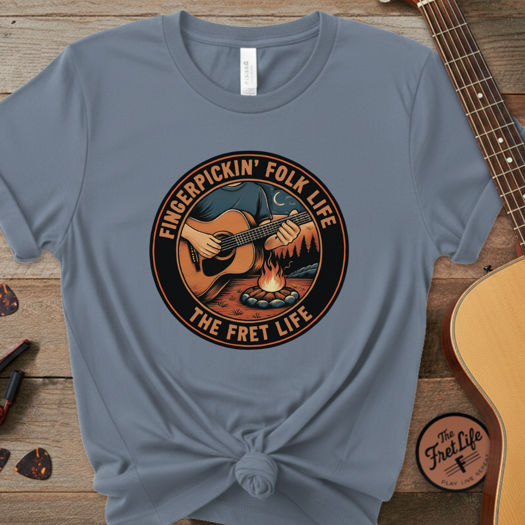 Blue t-shirt with 'Fingerpickin' Folk Life' graphic on a wooden surface with a guitar.