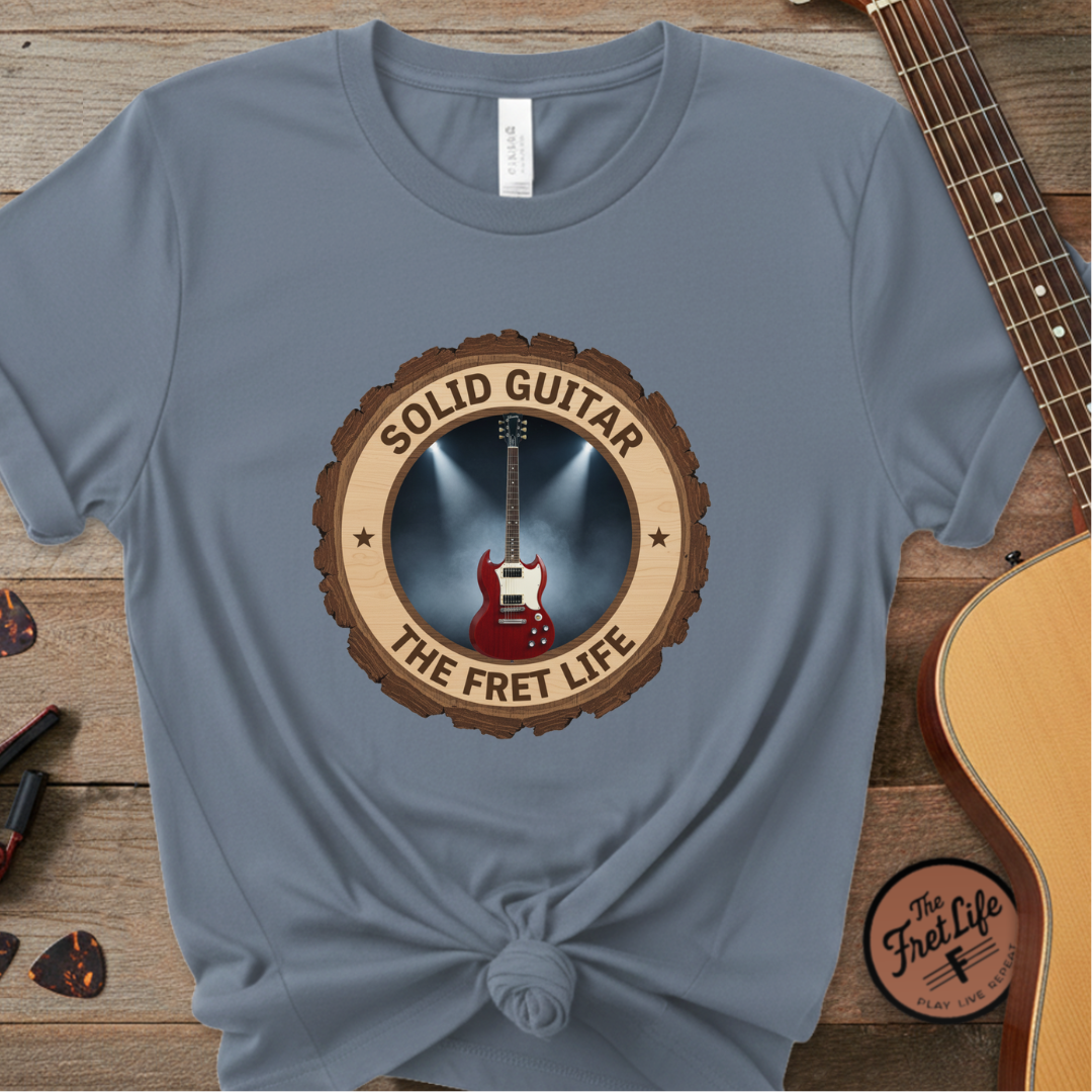 Blue t-shirt with guitar graphic and 'Solid Guitar The Fret Life' text on a wooden surface with guitars.