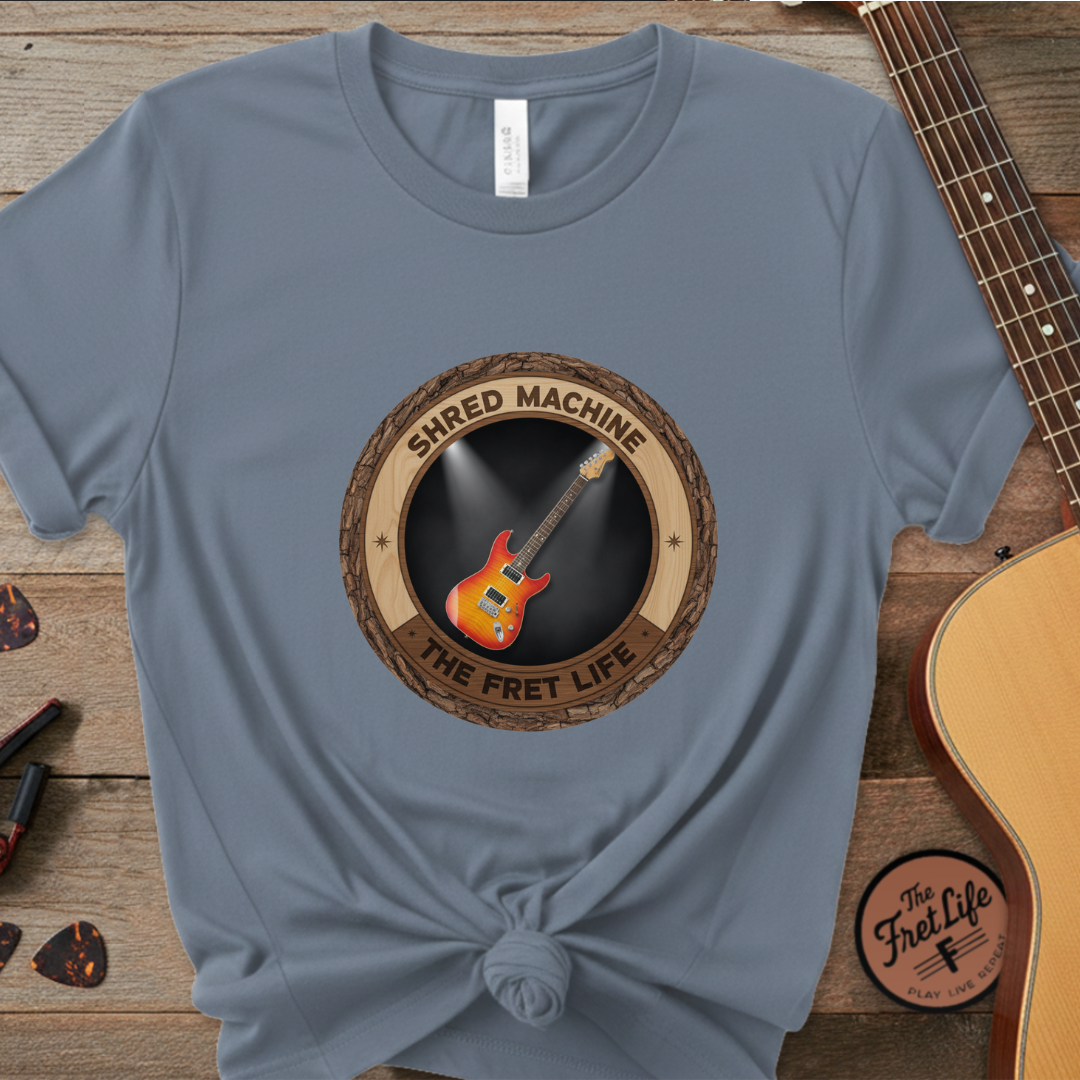 Blue t-shirt with 'Shred Machine' graphic on a wooden surface with guitars.