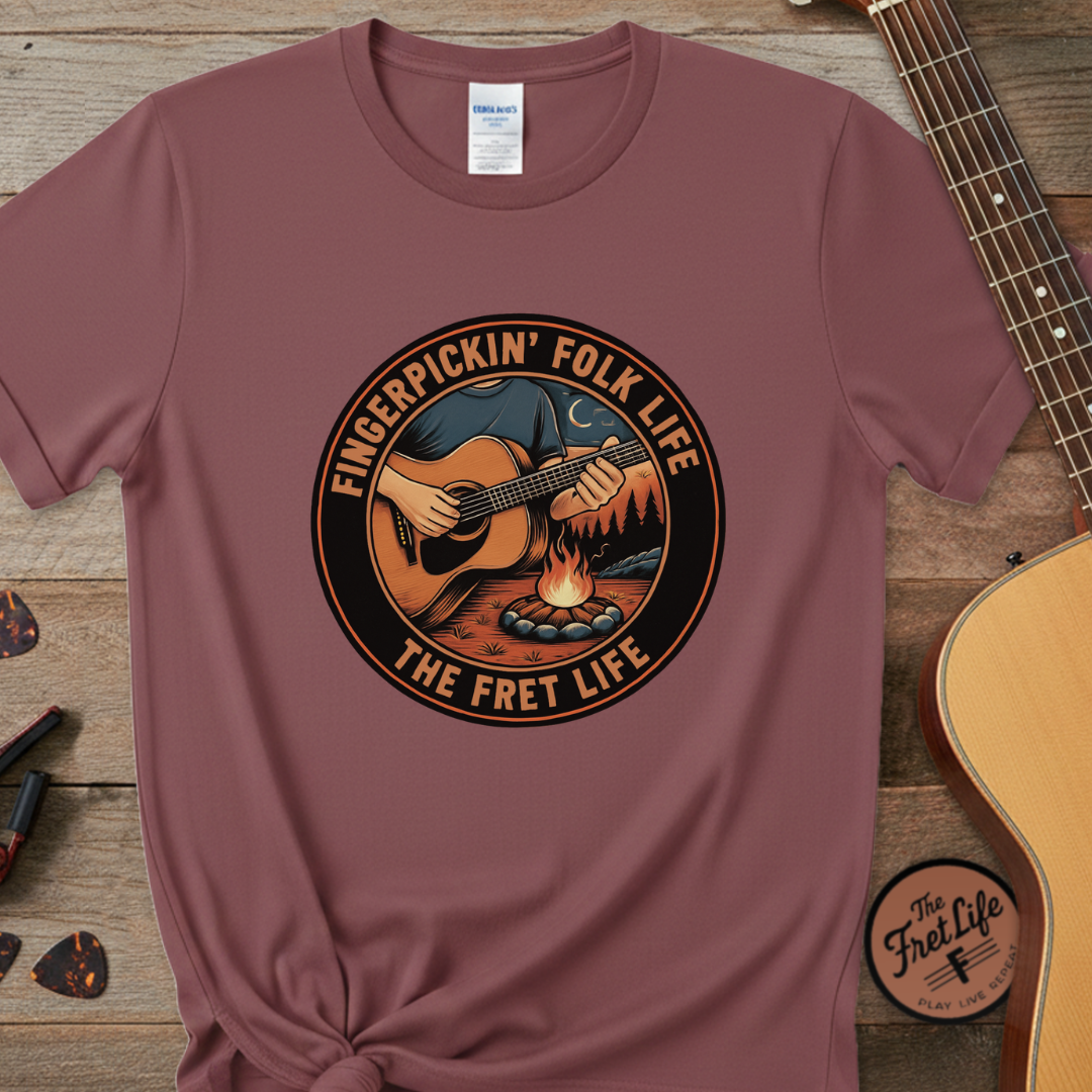 Maroon t-shirt with 'Fingerpickin' Folk Life' graphic on a wooden surface with a guitar.