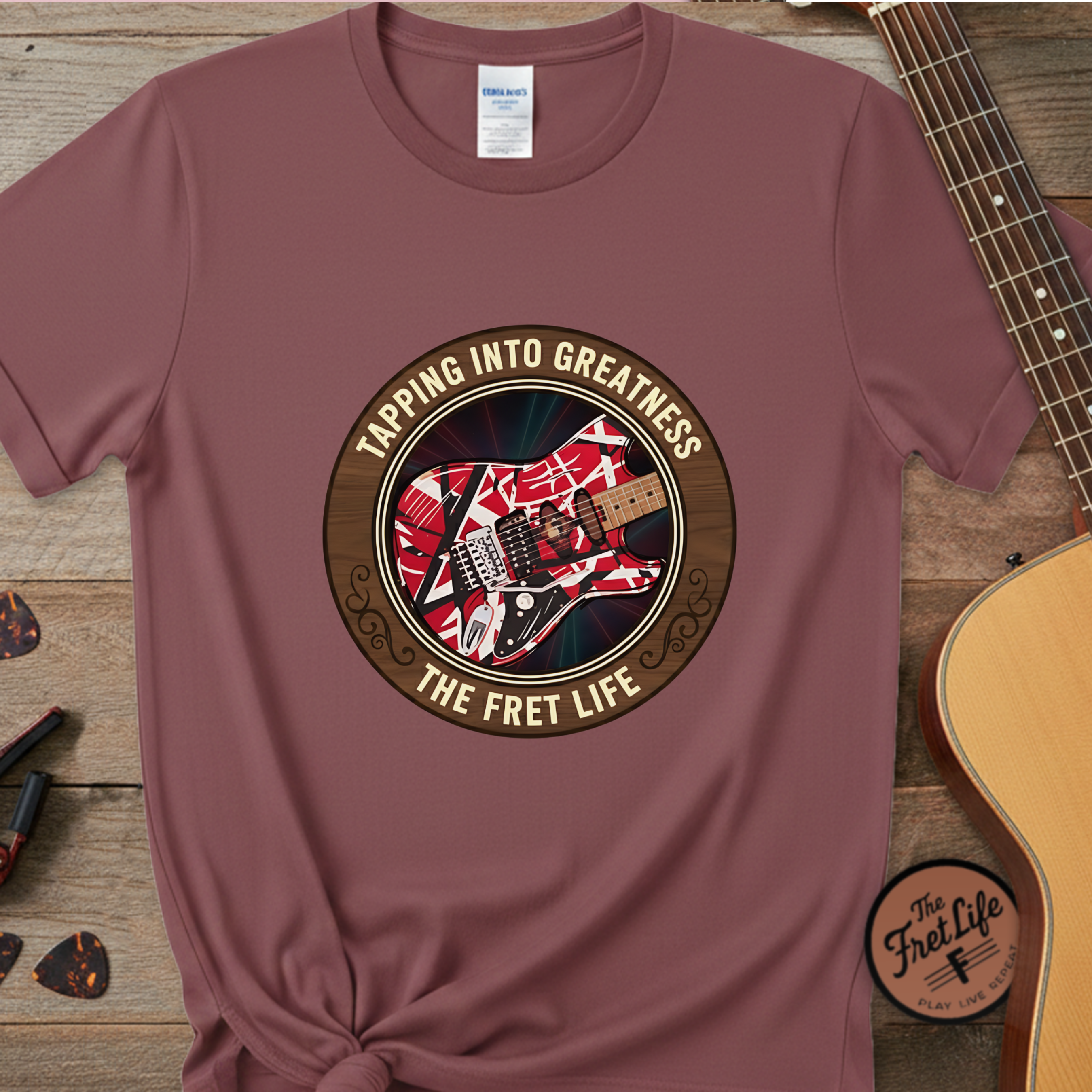 Maroon t-shirt with guitar graphic and text 'Tapping into Greatness, The Fret Life' on a wooden surface with a guitar.