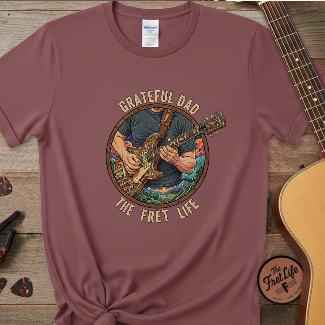 Maroon t-shirt with 'Grateful Dad The Fret Life' graphic on a wooden surface with a guitar.