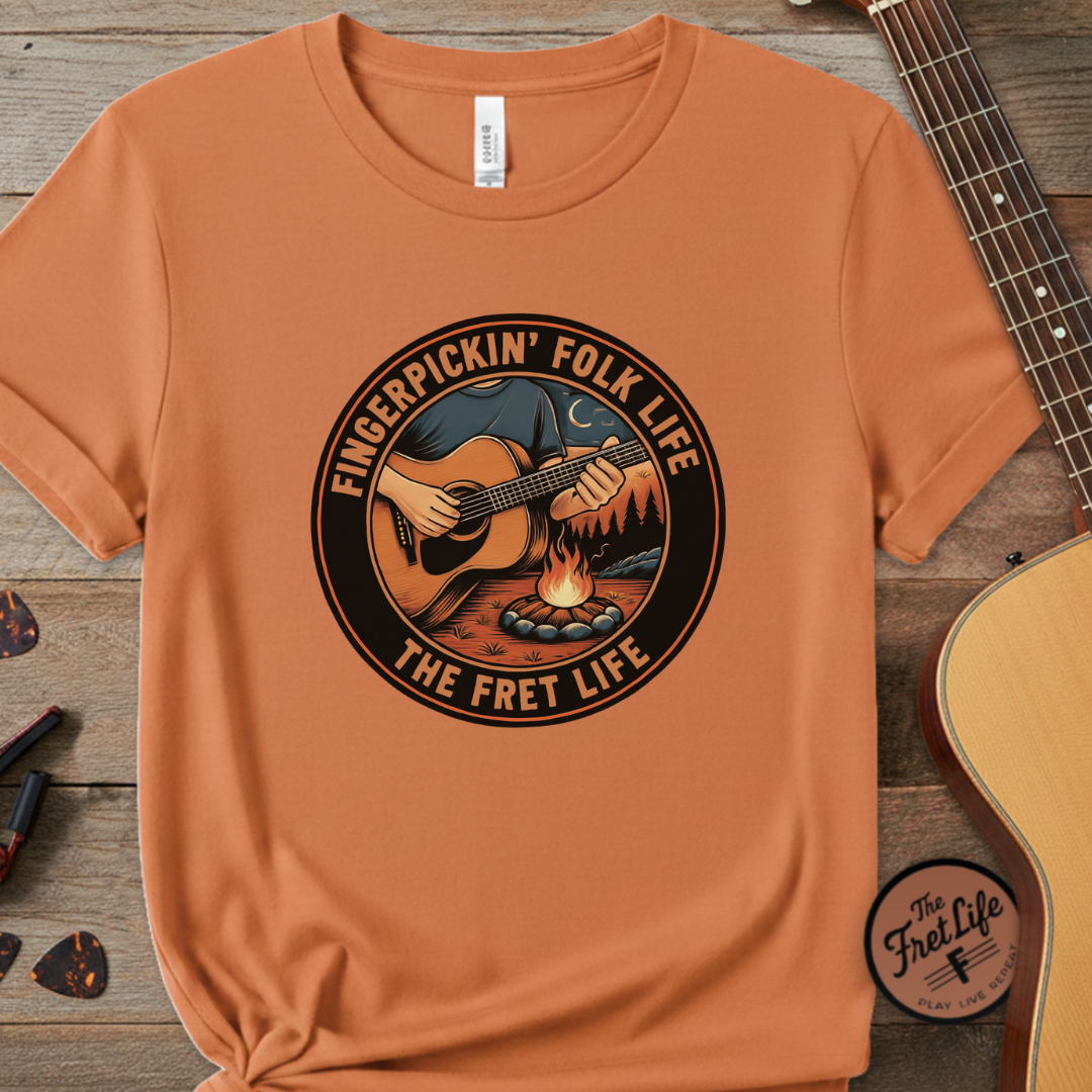 Orange t-shirt with 'Fingerpickin' Folk Life' graphic on a wooden surface with a guitar.