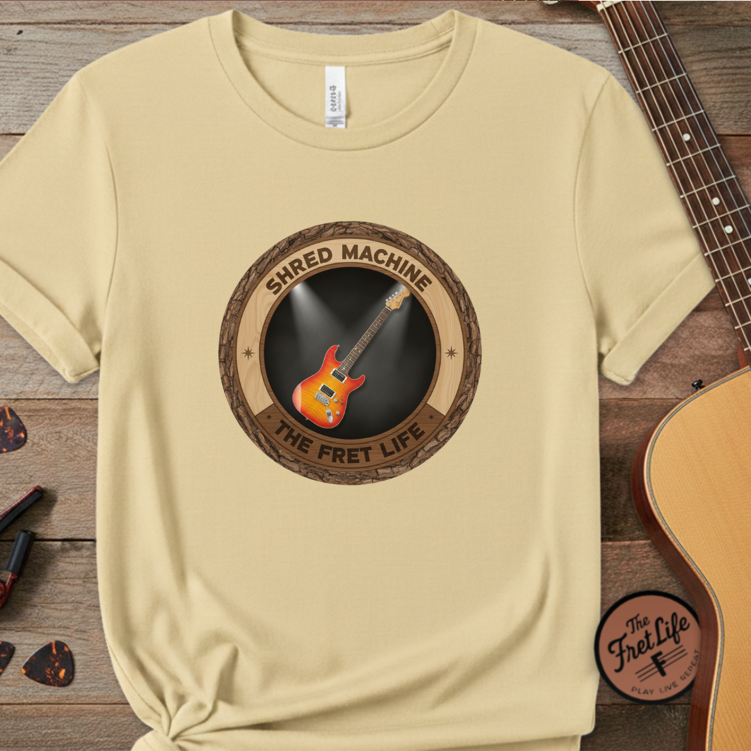 Beige t-shirt with 'Shred Machine' graphic on a wooden surface with a guitar