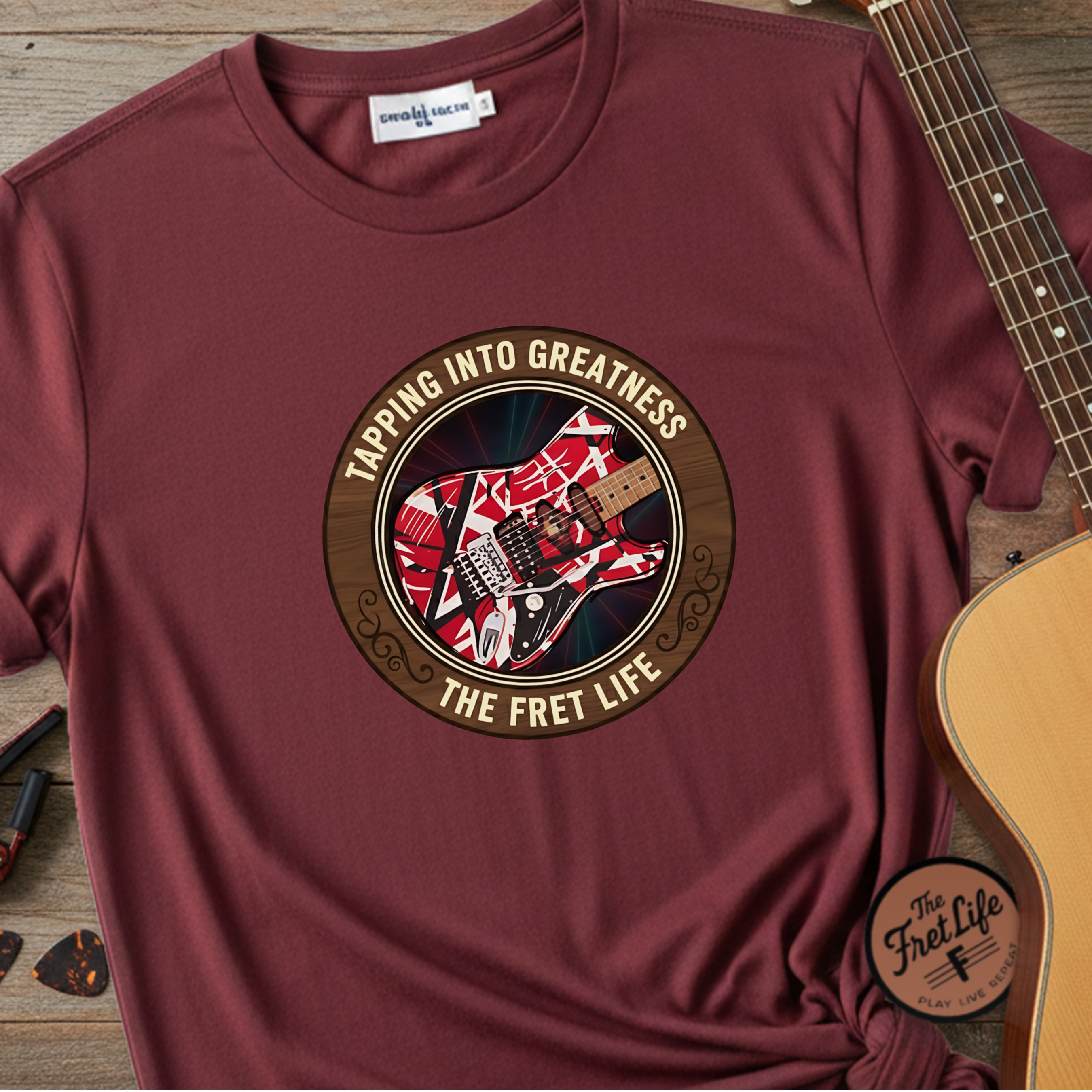 Maroon t-shirt with guitar graphic and text 'Tapping Into Greatness' on a wooden surface with a guitar.