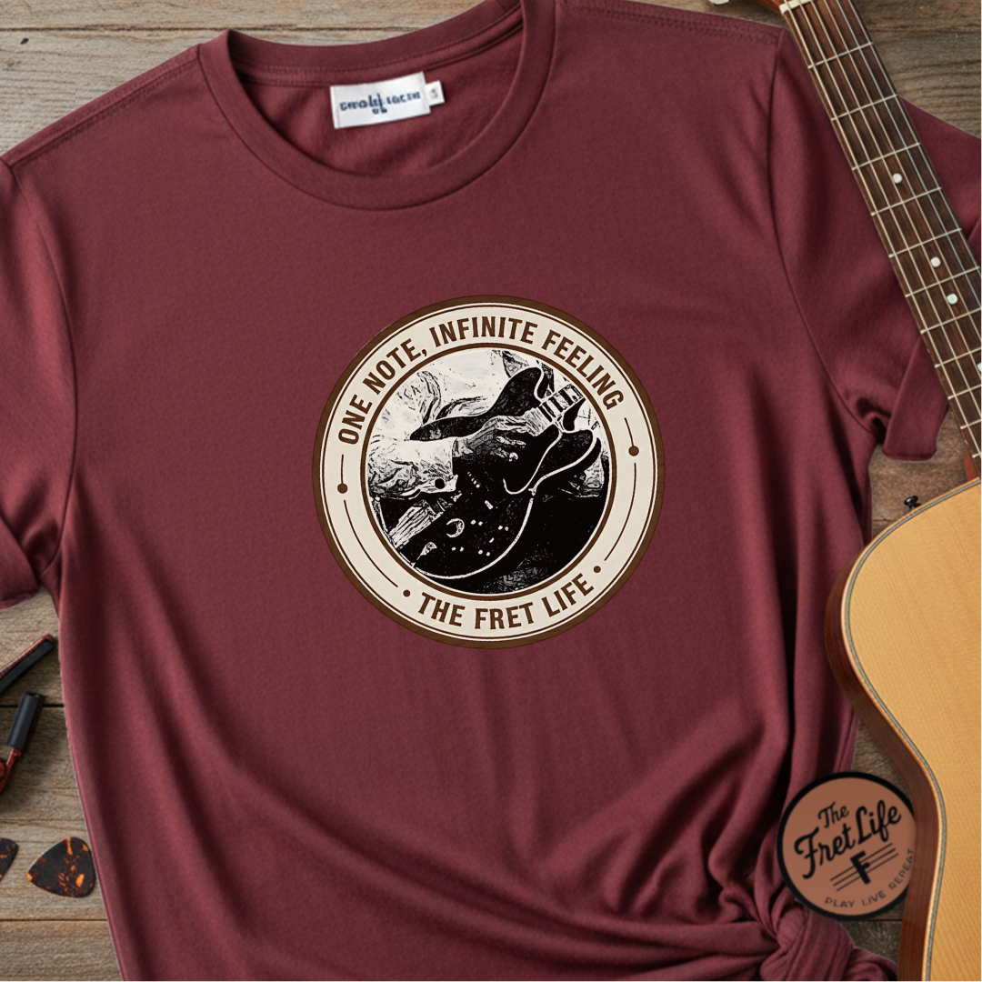 Maroon t-shirt with 'The Fret Life' graphic design on a wooden surface with a guitar.