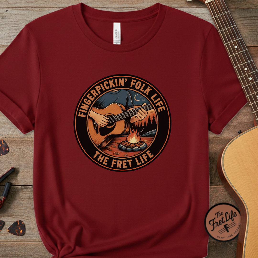 Maroon t-shirt with 'Fingerpickin' Folk Life' design on a wooden surface with a guitar.