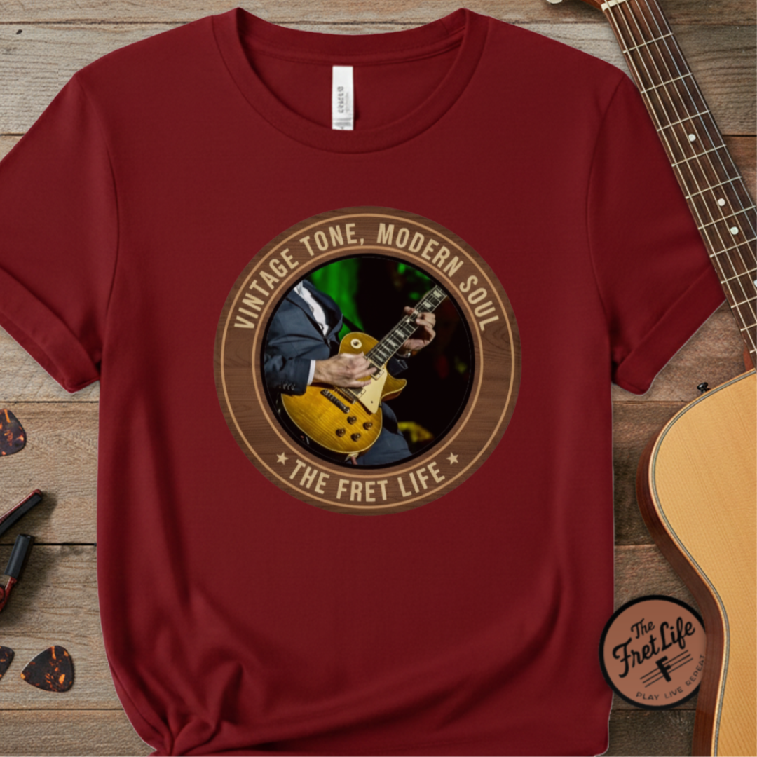 Red t-shirt with a graphic design of a person playing guitar and text 'Vintage Tone, Modern Soul The Fret Life' on a wooden surface with a guitar.