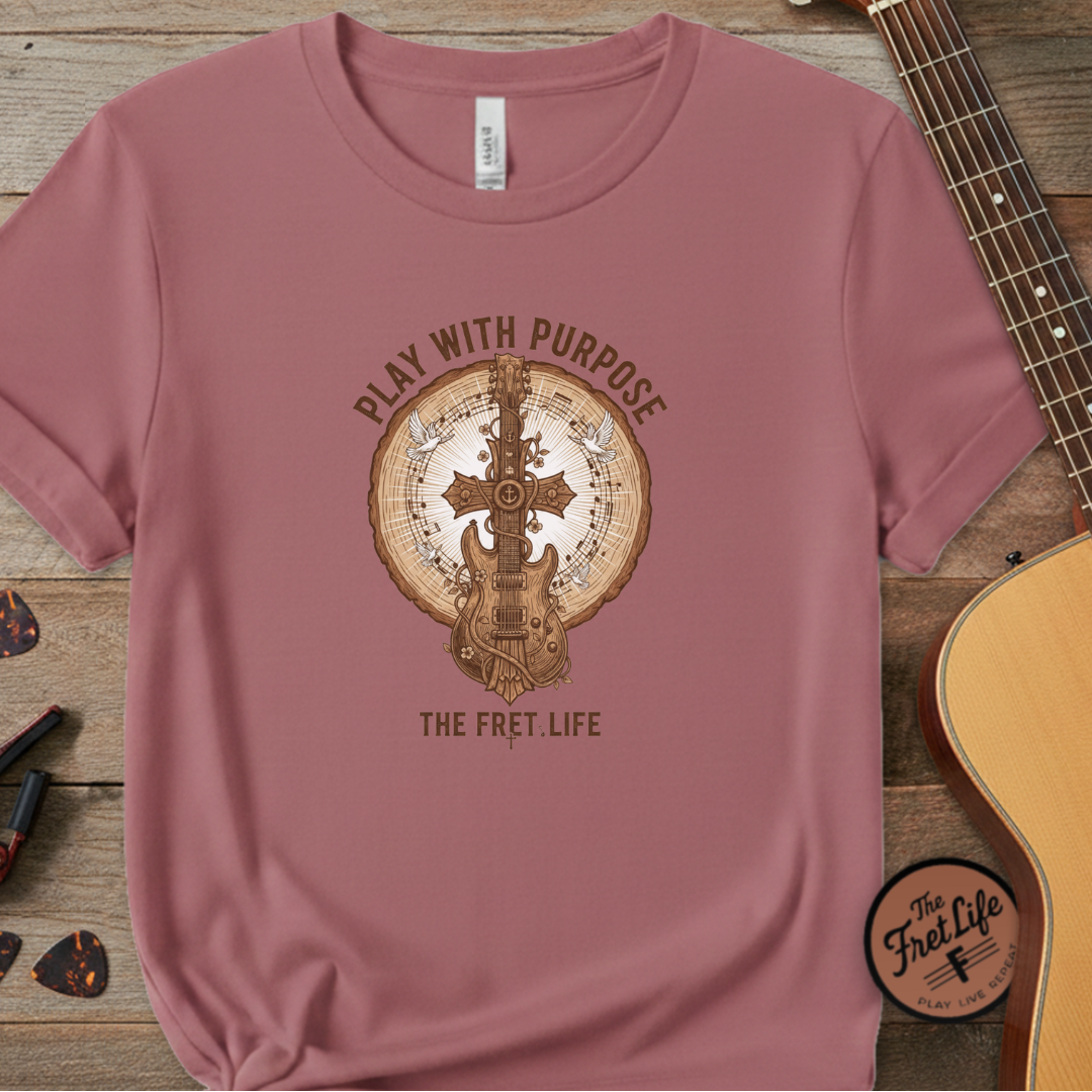 Pink t-shirt with guitar graphic and text 'Play with Purpose, The Fret Life' on a wooden surface with a guitar.