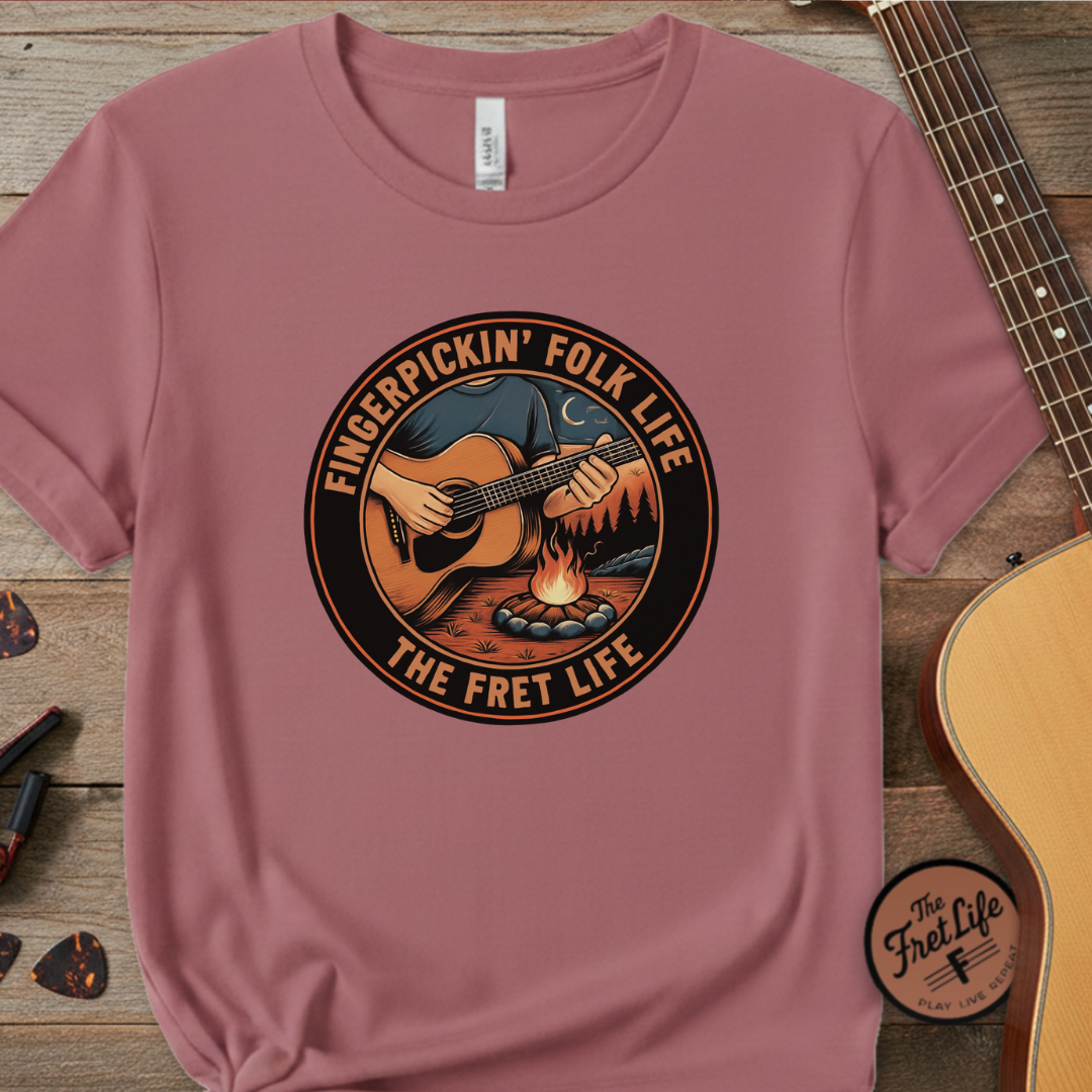 Pink t-shirt with 'Fingerpickin' Folk Life' graphic on a wooden surface with a guitar.