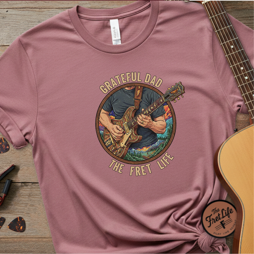 Mauve t-shirt with 'Grateful Dad The Fret Life' graphic on a wooden surface with a guitar.