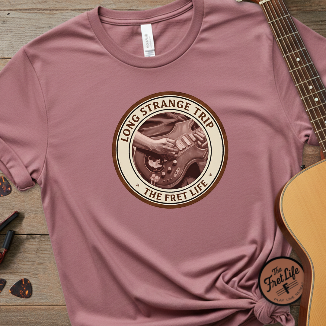 Pink t-shirt with 'Long Strange Trip' graphic design on a wooden surface with a guitar.