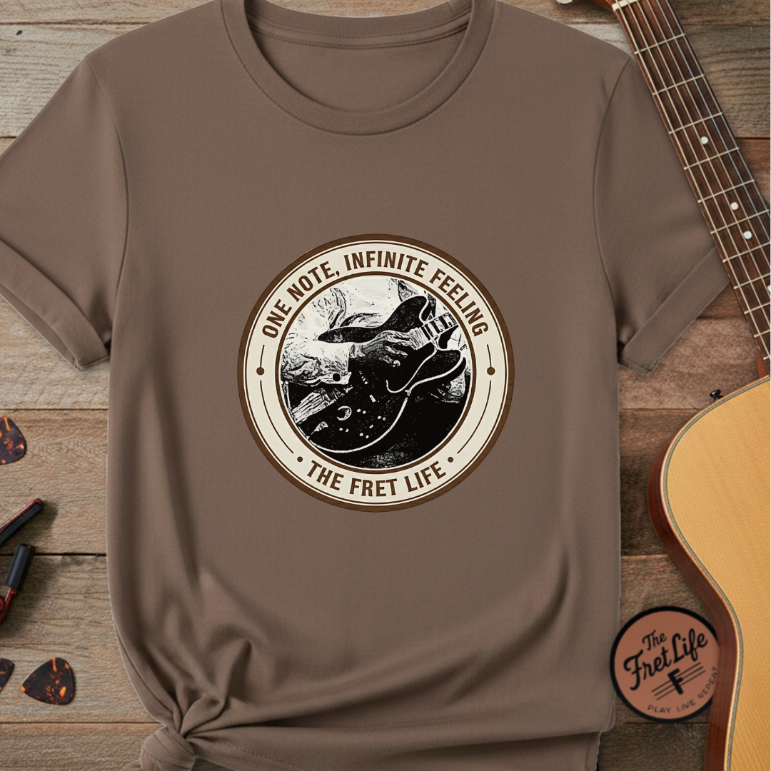 Brown t-shirt with 'The Fret Life' graphic design on a wooden surface with a guitar.