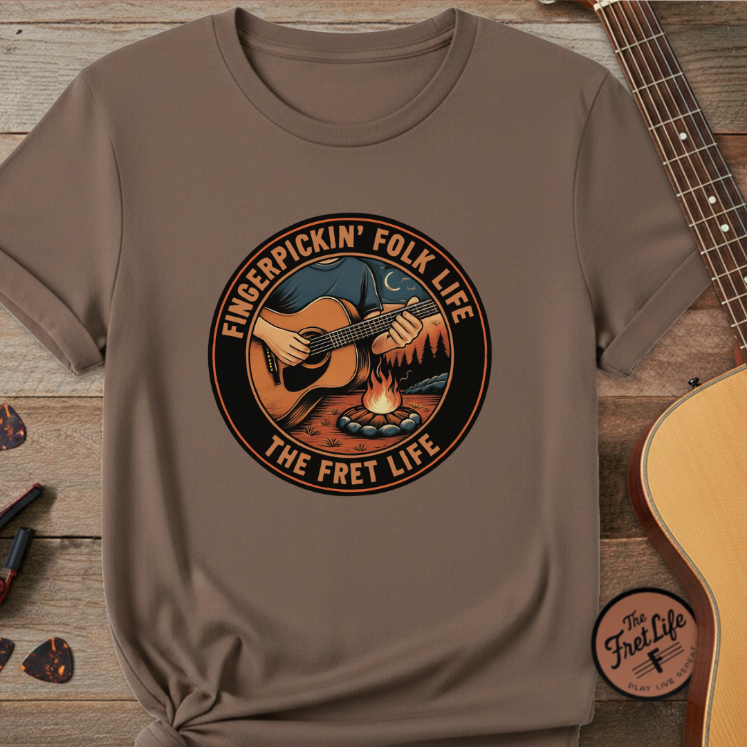 Brown t-shirt with 'Fingerpickin' Folk Life The Fret Life' graphic on a wooden surface with a guitar.