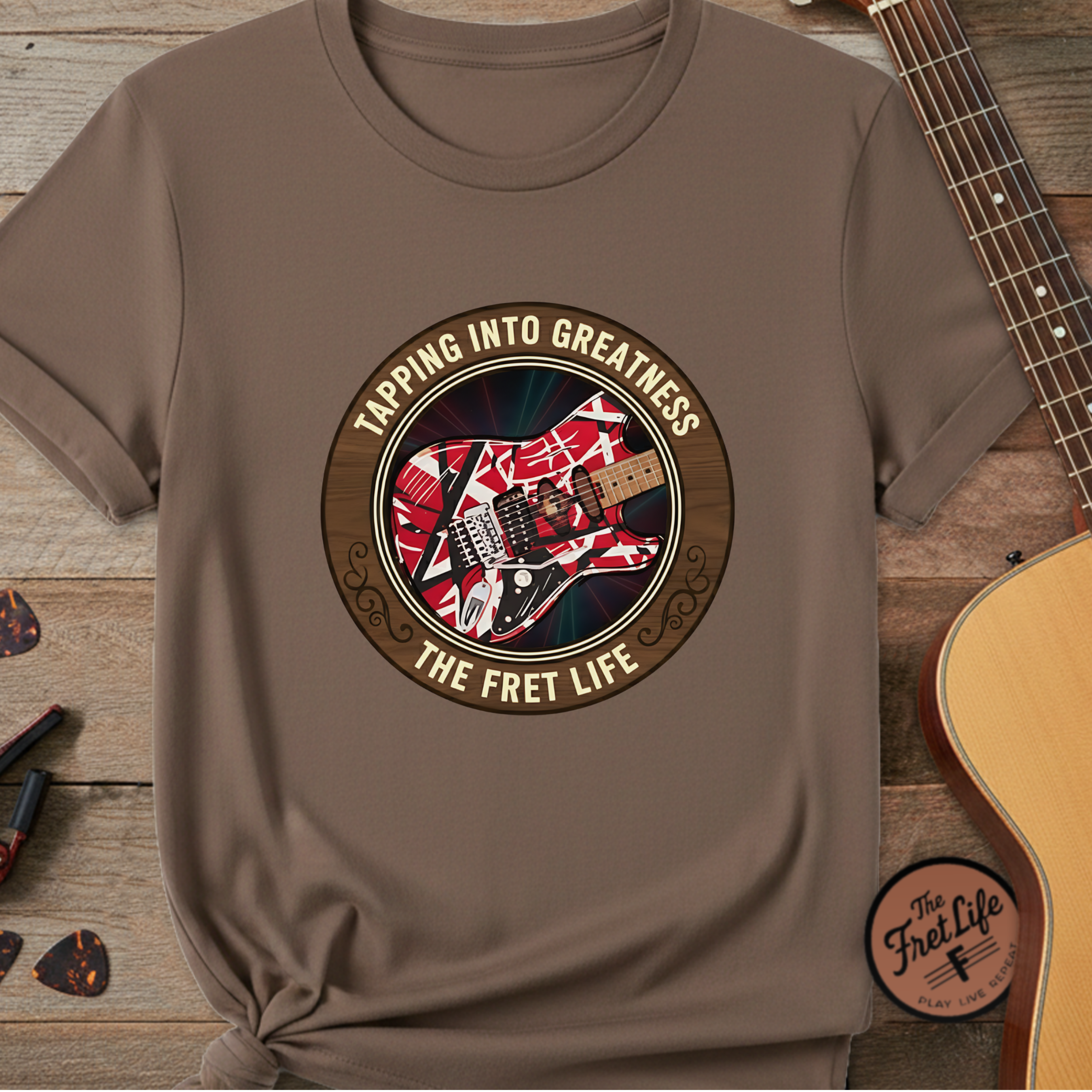 Brown t-shirt with guitar graphic and text 'Tapping into Greatness, The Fret Life' on a wooden surface with guitars.