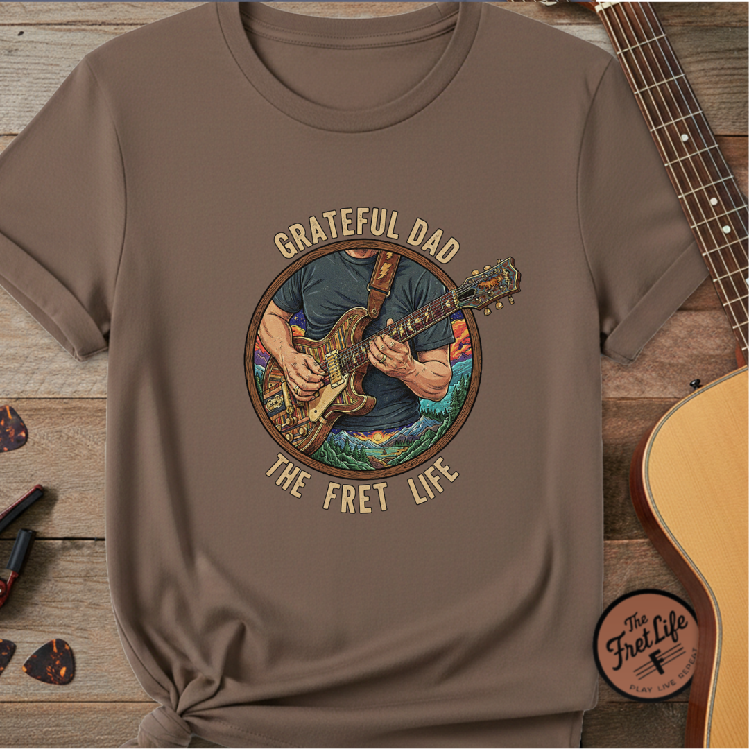 Brown t-shirt with 'Grateful Dad The Fret Life' graphic on a wooden surface with a guitar.