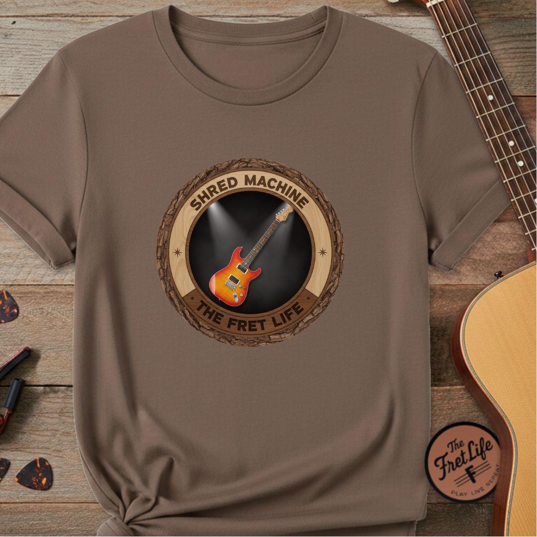 Brown t-shirt with guitar graphic and 'Shred Machine' text on a wooden surface with a guitar.