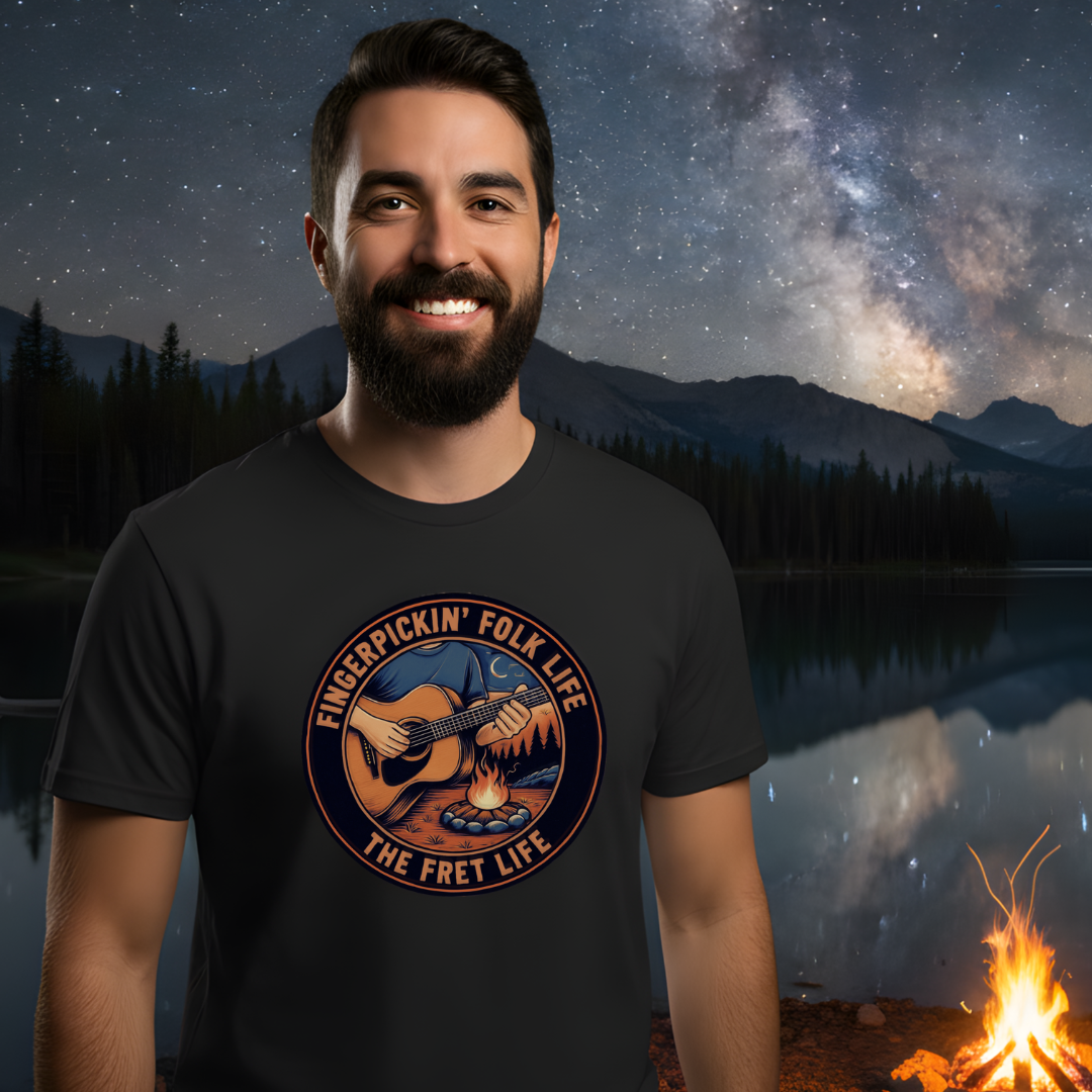 Man wearing a t-shirt with 'Fingerpickin' Folk Life' design against a scenic background with mountains and stars.