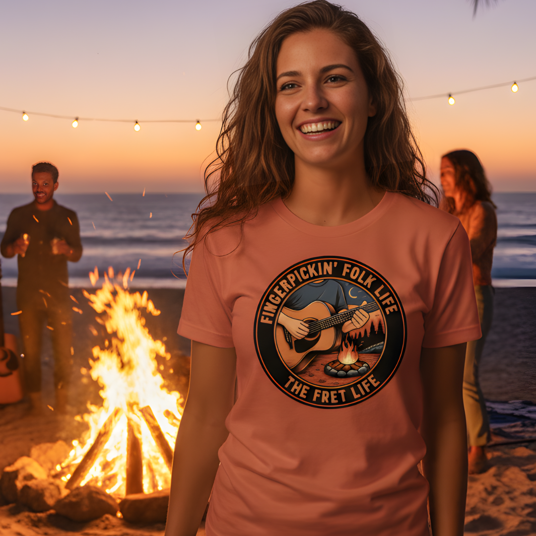Woman wearing a 'Fingerpicking Folk Life' t-shirt by a campfire on a beach at sunset.