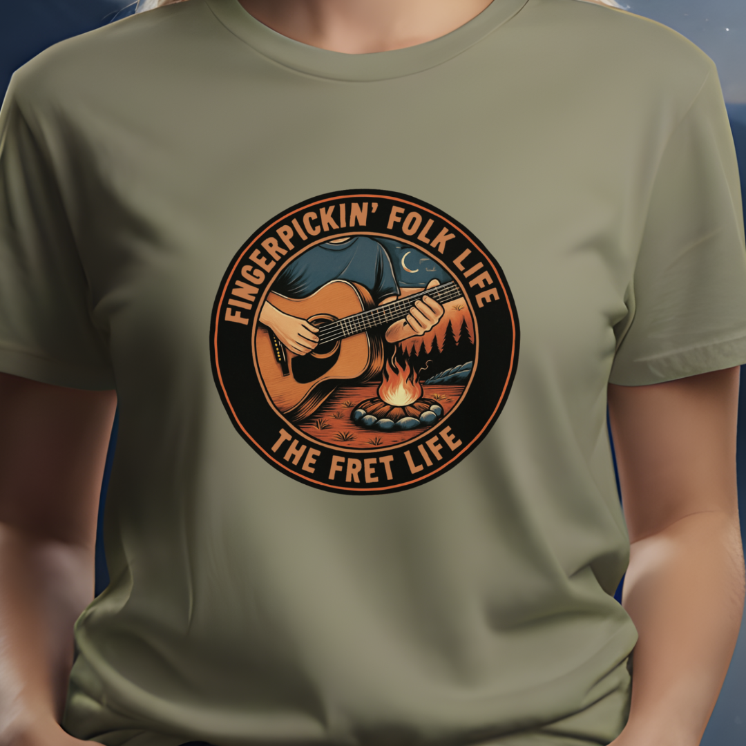 Person wearing a green t-shirt with a graphic design of a person playing guitar by a campfire and text 'Fingerpickin' Folk Life The Fret Life'.