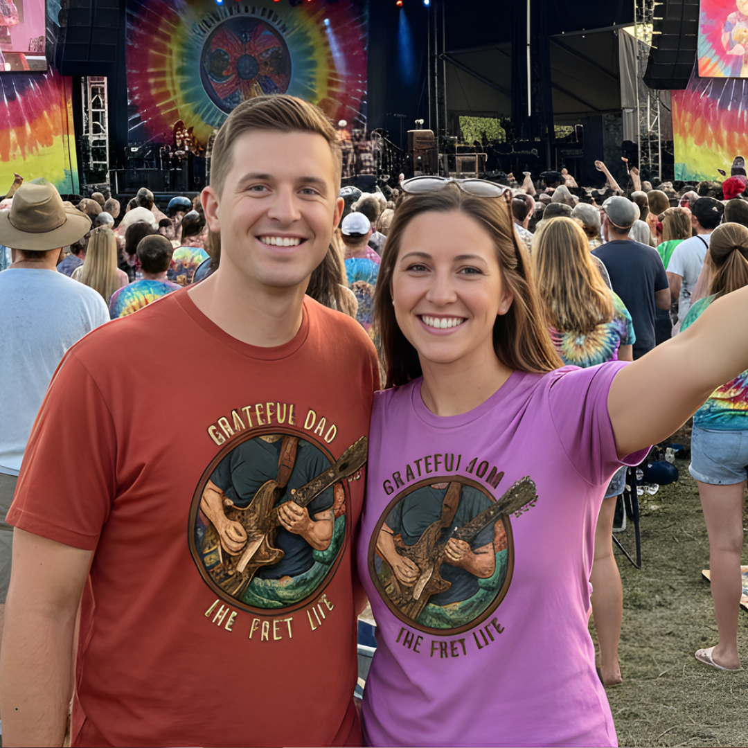Two people wearing 'Grateful Dad' t-shirts at a music festival with a crowd and colorful stage in the background.