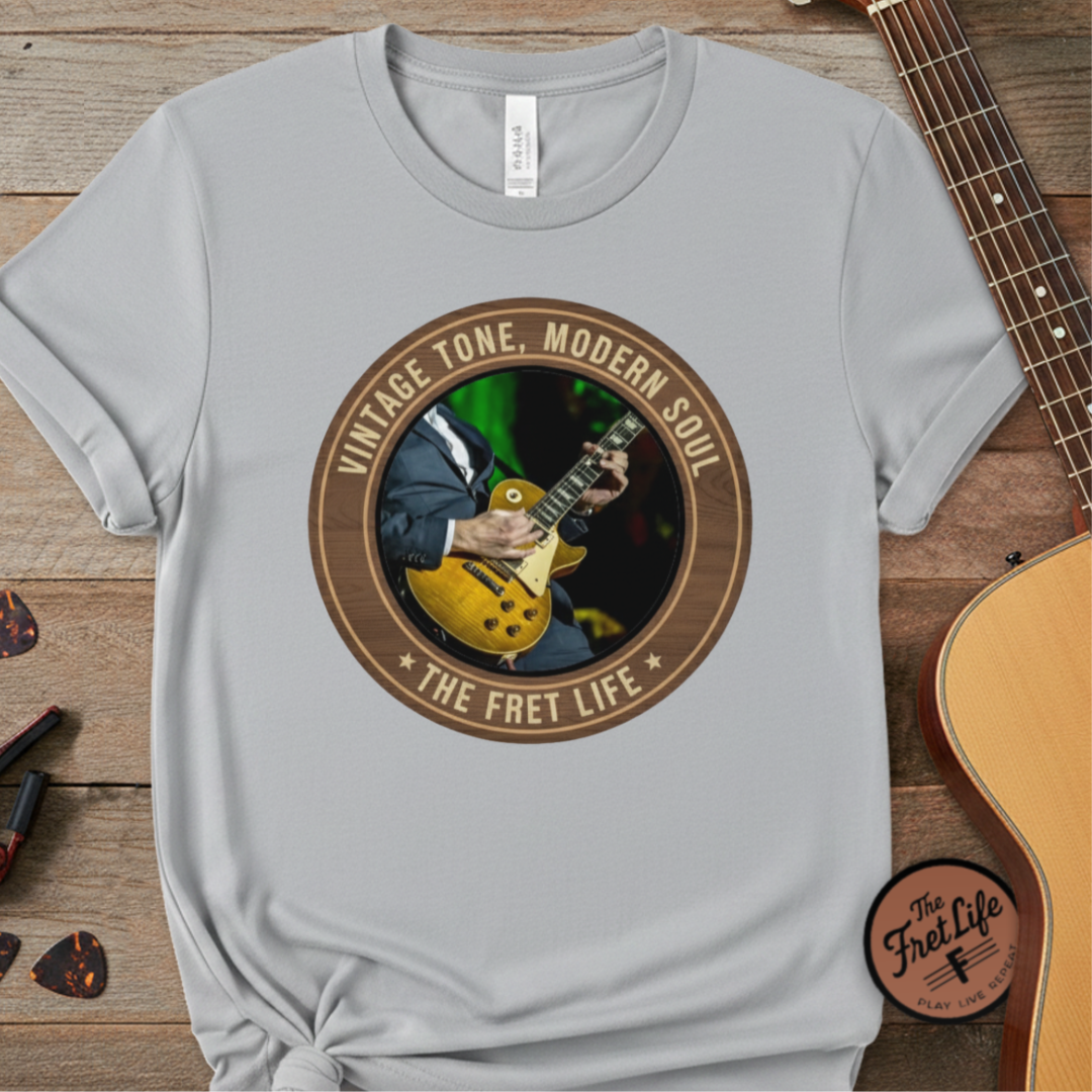Gray t-shirt with a guitar graphic and text on a wooden surface with a guitar.