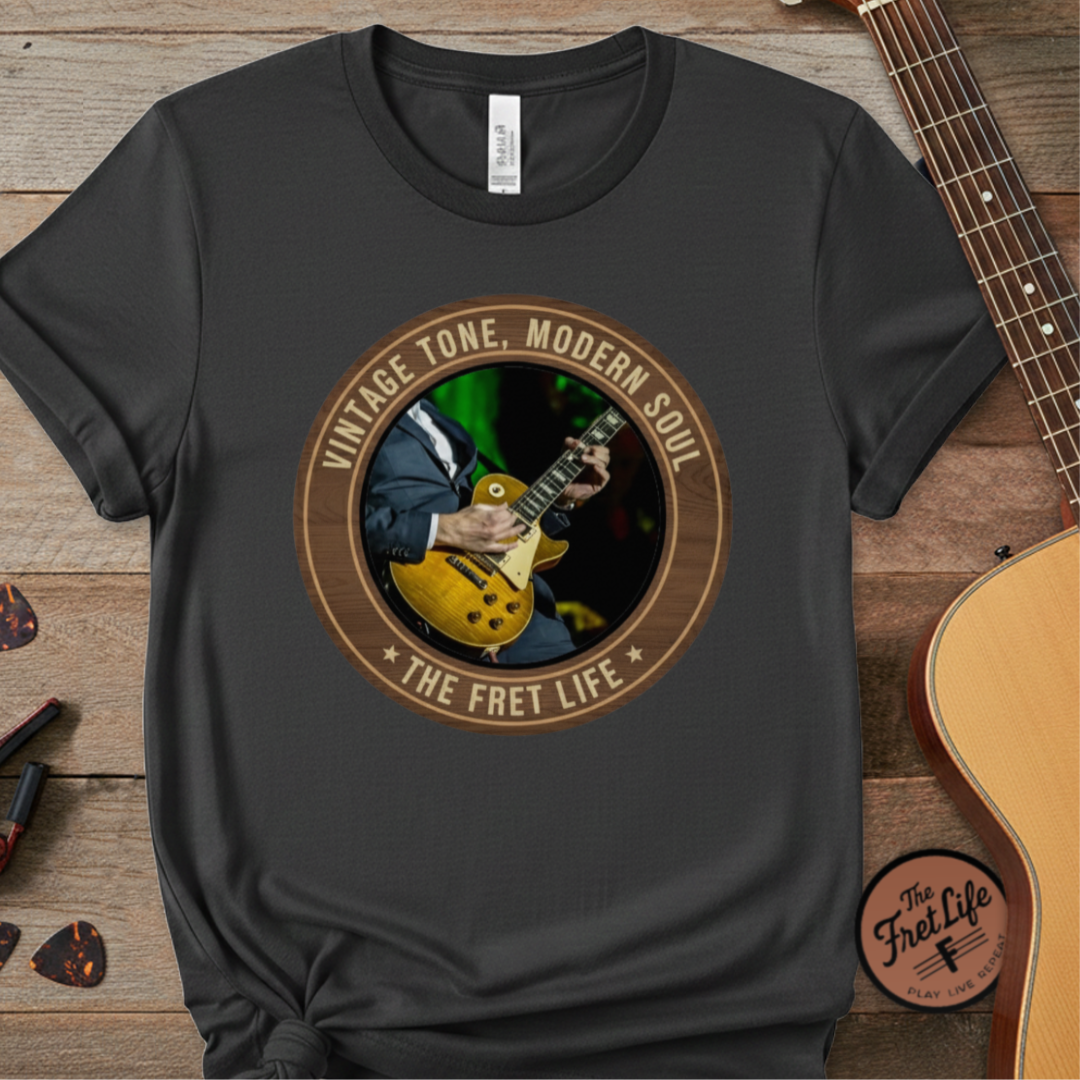 Black t-shirt with guitar graphic and 'The Fret Life' text on a wooden surface with a guitar.