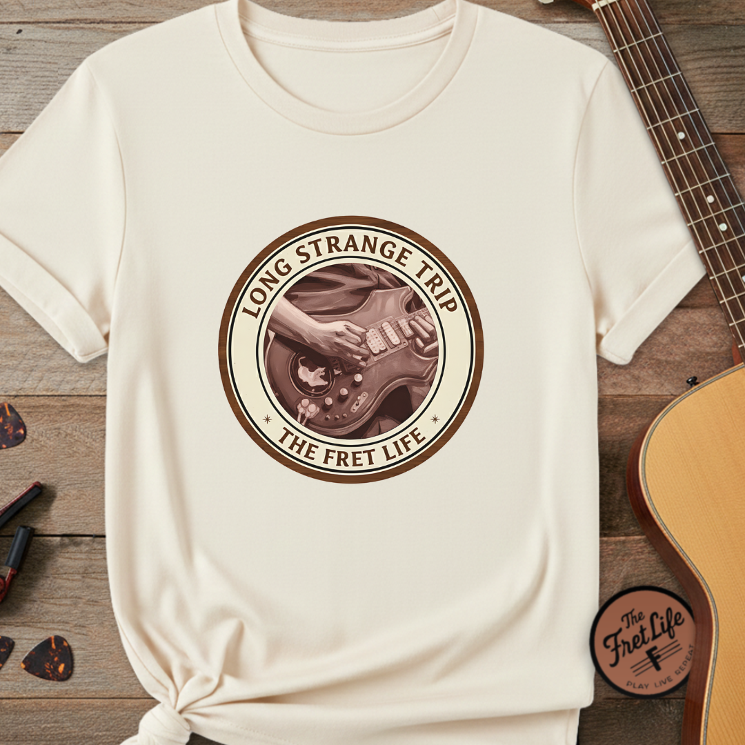 White t-shirt with 'Long Strange Trip: The Fret Life' graphic on a wooden surface with a guitar and pickguard.