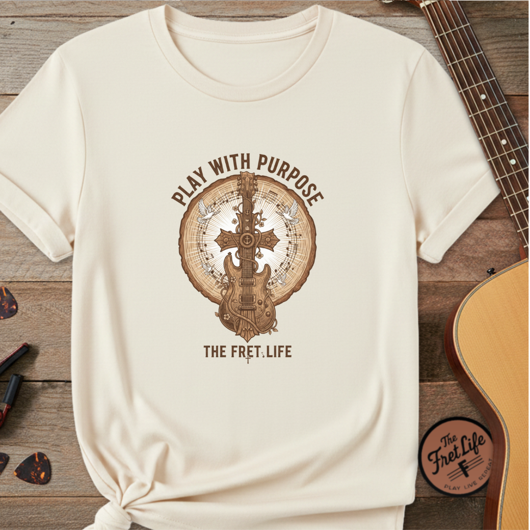 Beige t-shirt with guitar graphic and 'Play with Purpose' text on a wooden surface with a guitar.