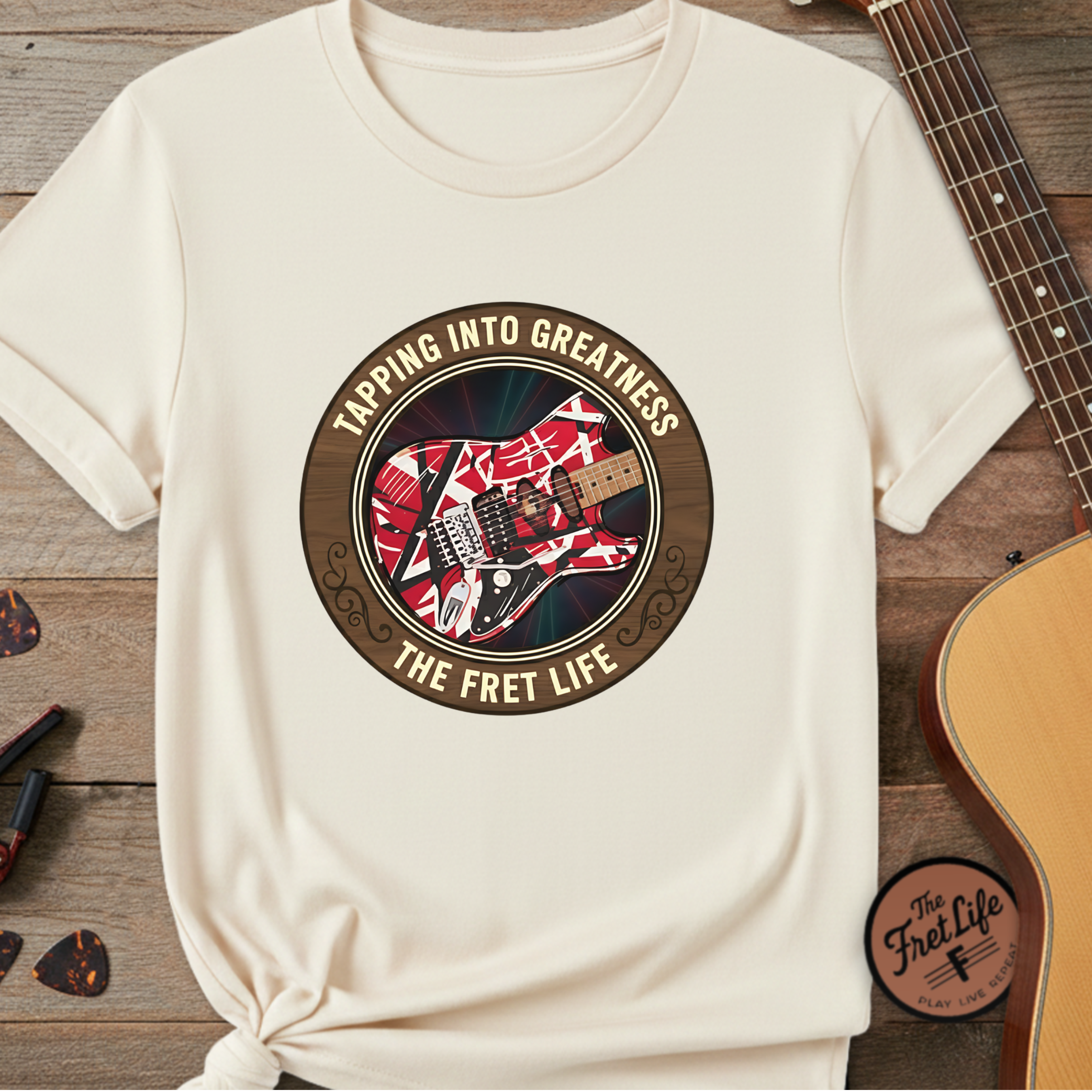 White t-shirt with guitar graphic and text 'Tapping into Greatness, The Fret Life' on a wooden surface with a guitar.
