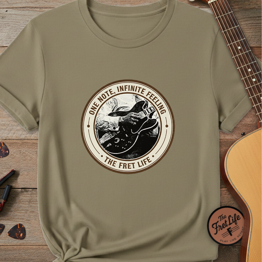 Khaki t-shirt with 'The Fret Life' graphic design on a wooden surface with a guitar.