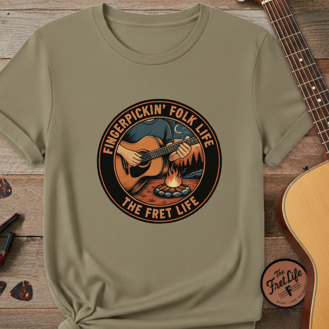 Khaki t-shirt with 'Fingerpickin' Folk Life, The Fret Life' graphic on a wooden surface with a guitar.
