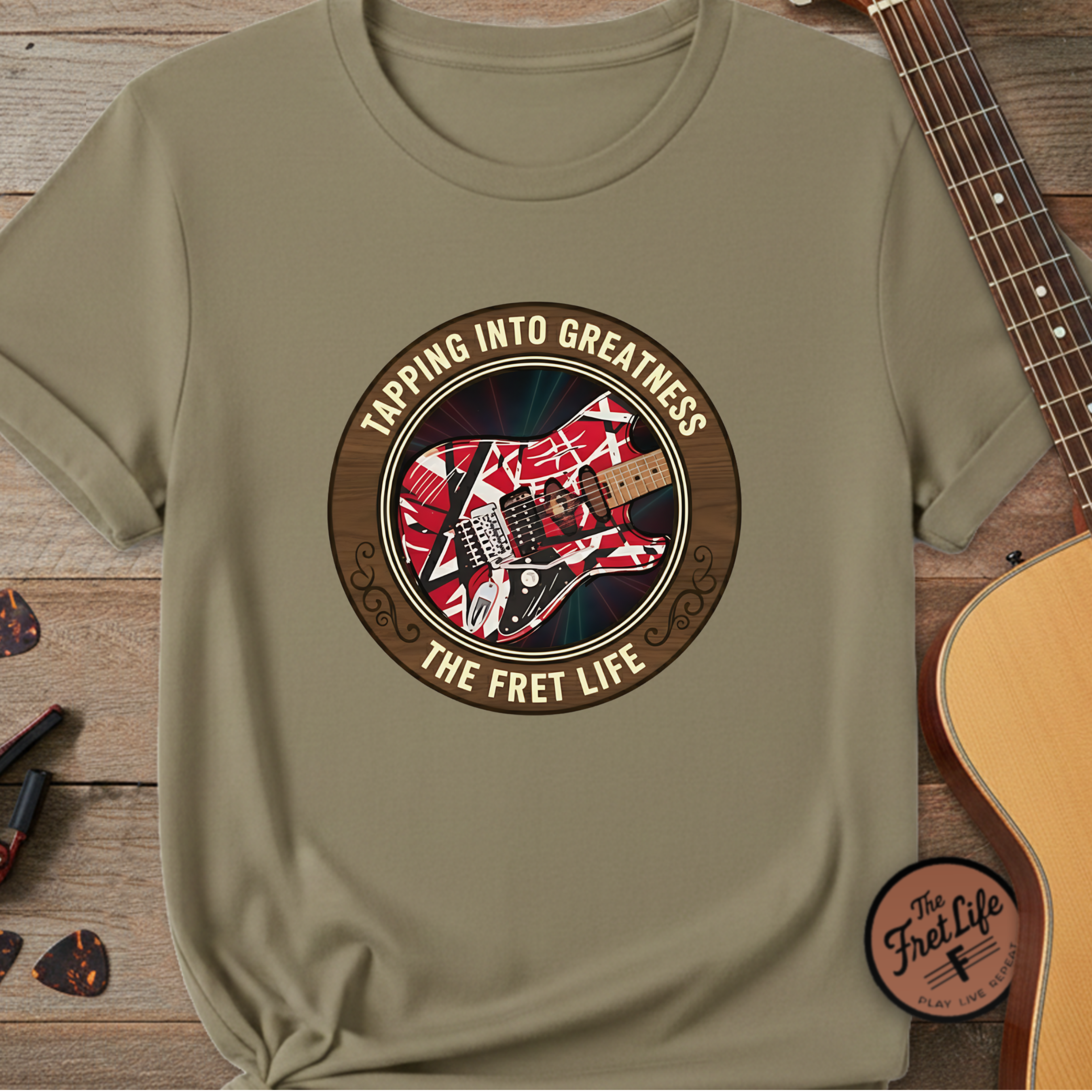 Khaki t-shirt with guitar graphic and text 'Tapping into Greatness, The Fret Life' on a wooden surface with a guitar.