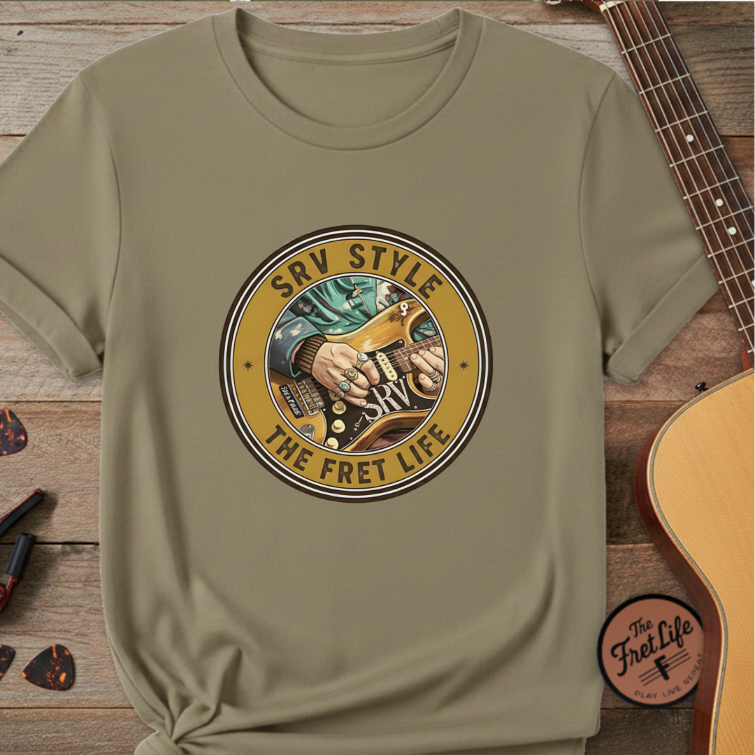 Khaki t-shirt with guitar graphic and 'SRV Style The Fret Life' text on a wooden surface with a guitar.