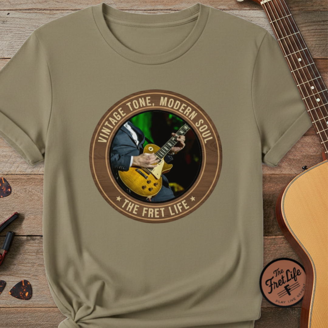 Khaki t-shirt with guitar graphic and 'The Fret Life' text on a wooden surface with a guitar.