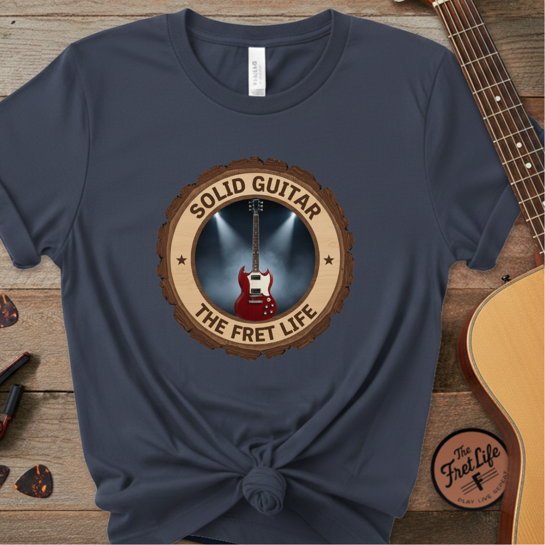 Navy blue t-shirt with 'Solid Guitar The Fret Life' design on a wooden background with guitars.