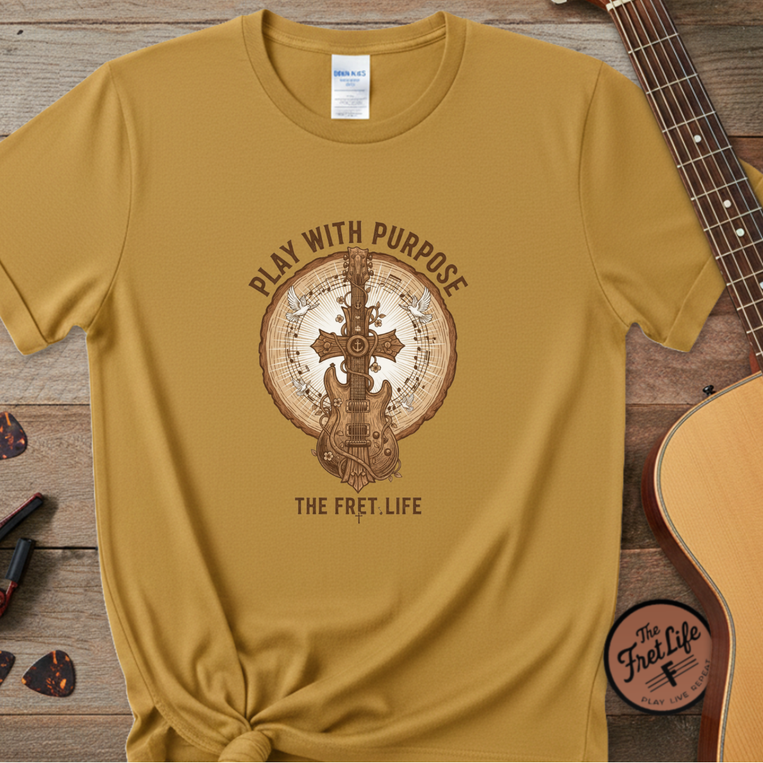 Mustard yellow t-shirt with guitar graphic and text 'Play with Purpose, The Fret Life' on a wooden surface with a guitar.