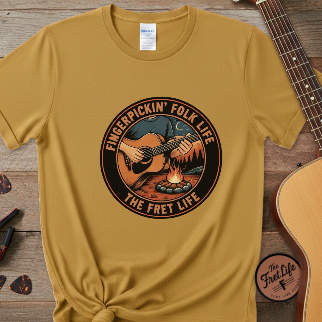 Mustard yellow t-shirt with 'Fingerpickin' Folk Life, The Fret Life' graphic on a wooden surface with a guitar.