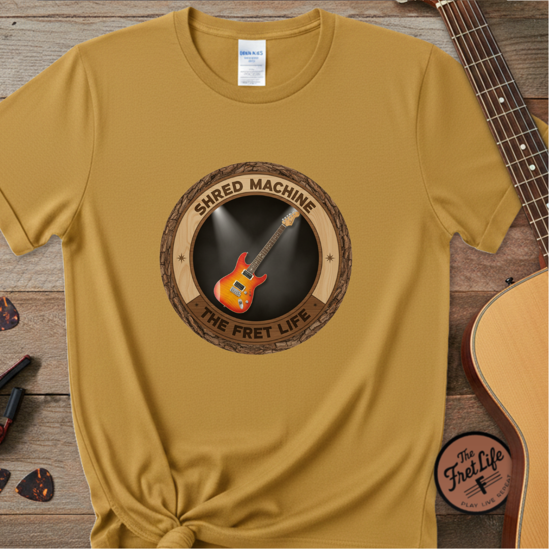 Mustard yellow t-shirt with 'Shred Machine' graphic on a wooden surface with a guitar.