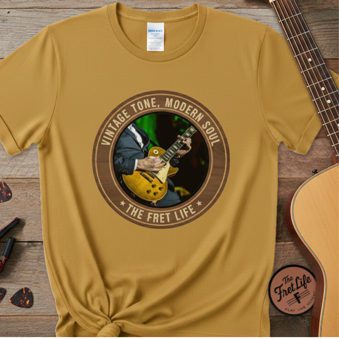 Yellow t-shirt with a graphic of a person playing guitar and text 'Vintage Tone, Modern Soul The Fret Life' on a wooden surface with a guitar.