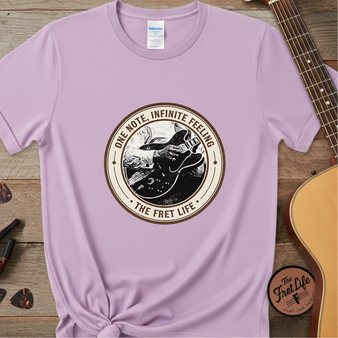 Light purple t-shirt with 'The Fret Life' graphic design on a wooden surface with a guitar.