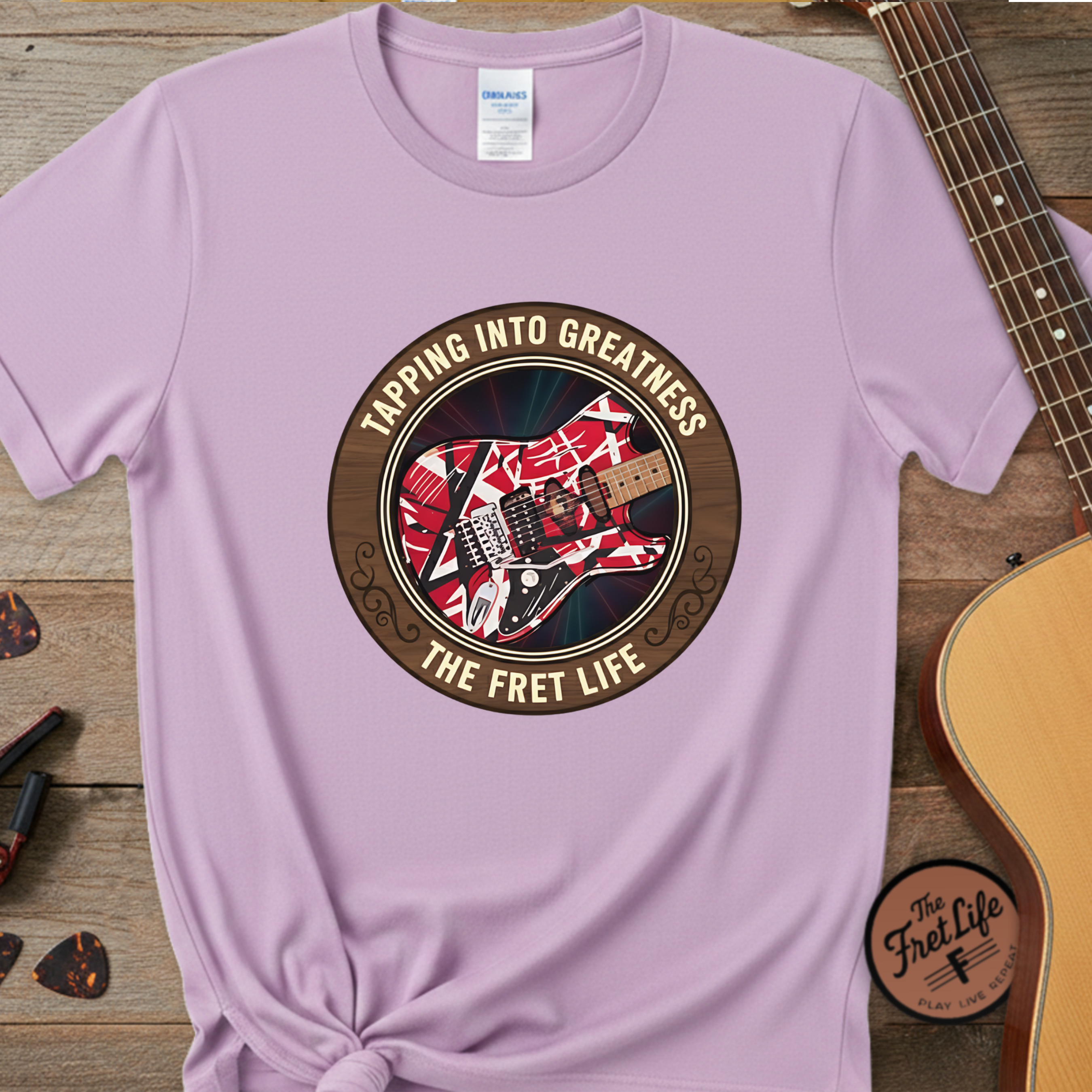 Purple t-shirt with guitar graphic and text 'Tapping Into Greatness, The Fret Life' on a wooden surface with a guitar.
