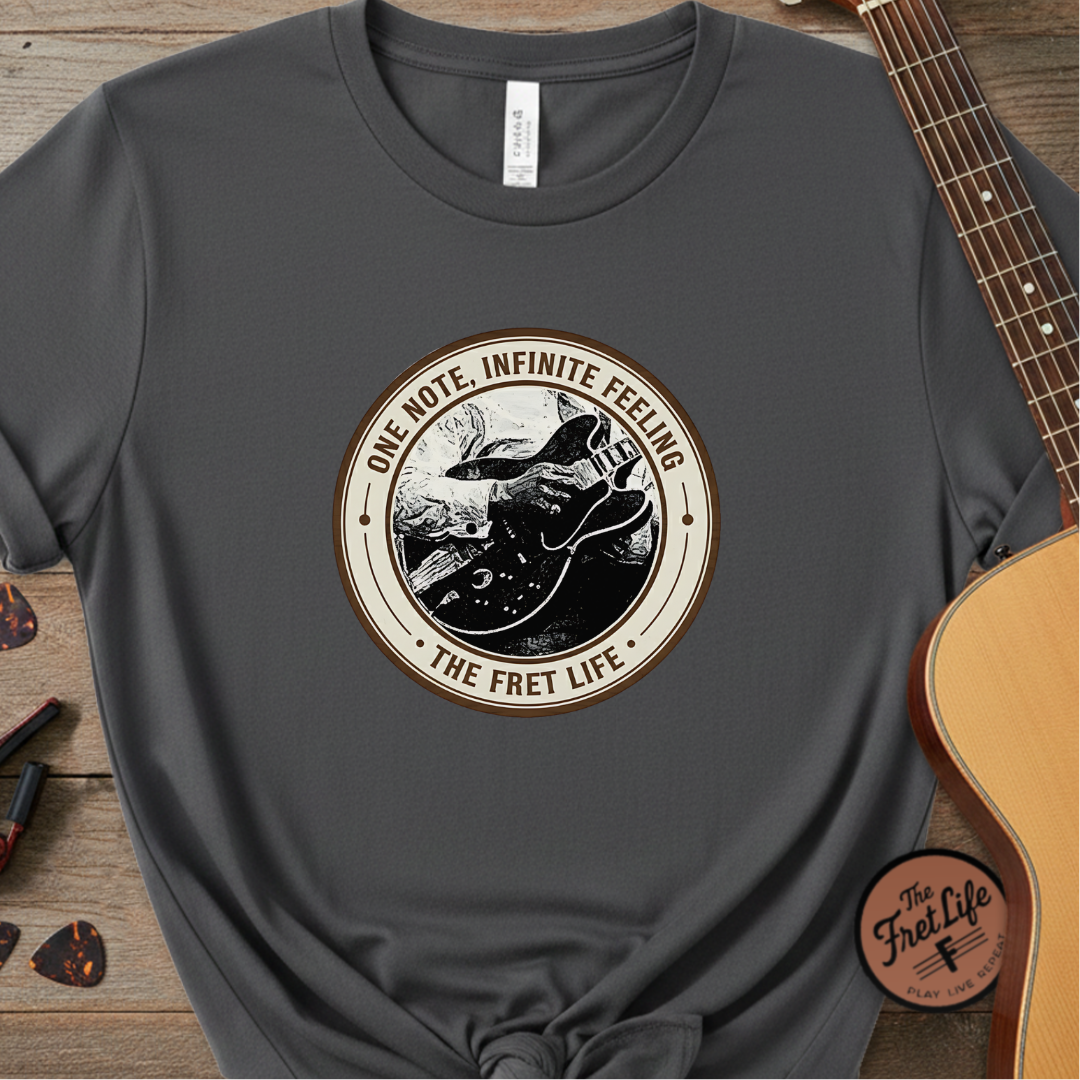 Gray t-shirt with 'The Fret Life' graphic design on a wooden surface with a guitar.