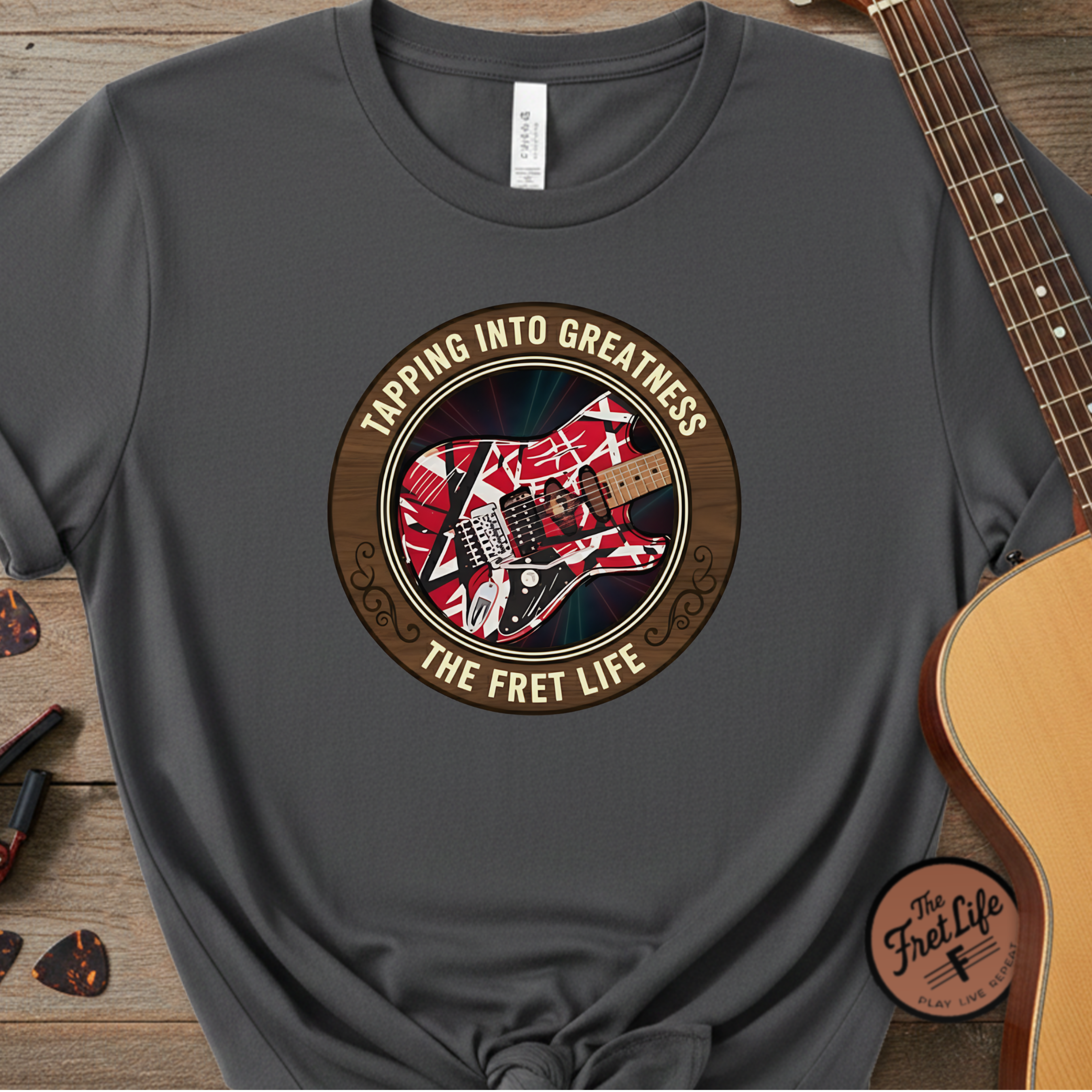 Gray t-shirt with guitar graphic and text 'Tapping into Greatness, The Fret Life' on a wooden surface with a guitar.